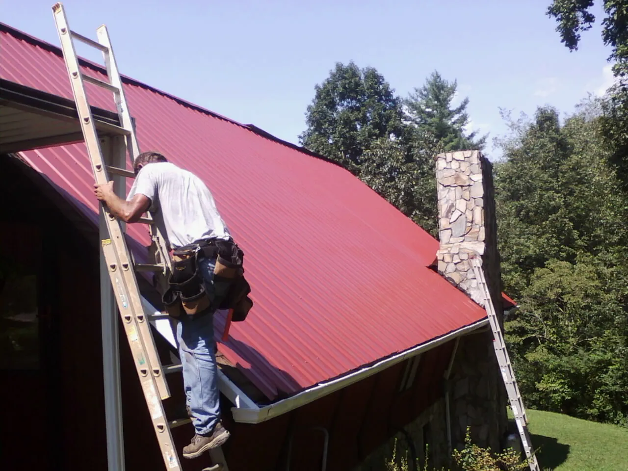 Roofer on a ladder beside a red metal roof and stone chimney — Soffit & Fascia Repair by Roofing Brothers in Weippe